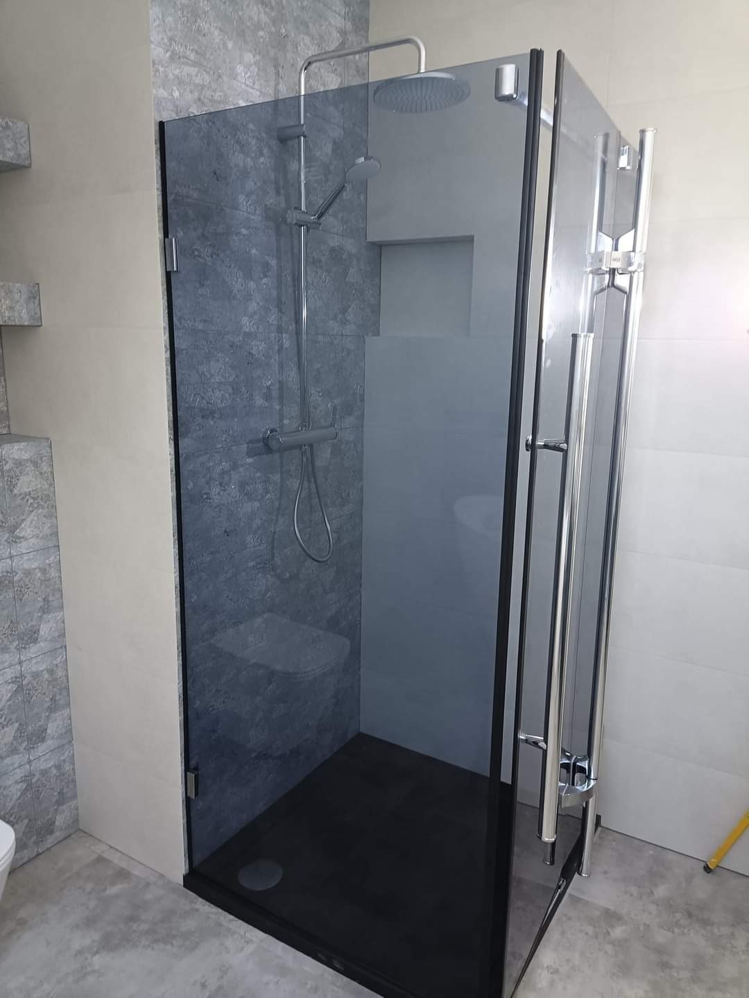 Kabina prysznicowa z grafitowym brodzikiem i szklanymi drzwiami, chromowana armatura, ściany wyłożone płytkami w odcieniach szarości i beżu, widoczny fragment toalety.