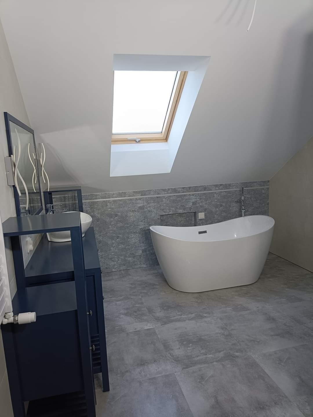 Nowoczesna łazienka na poddaszu z wolnostojącą białą wanną, umywalką nablatową na ciemnoniebieskiej szafce i oknem dachowym w skosie.