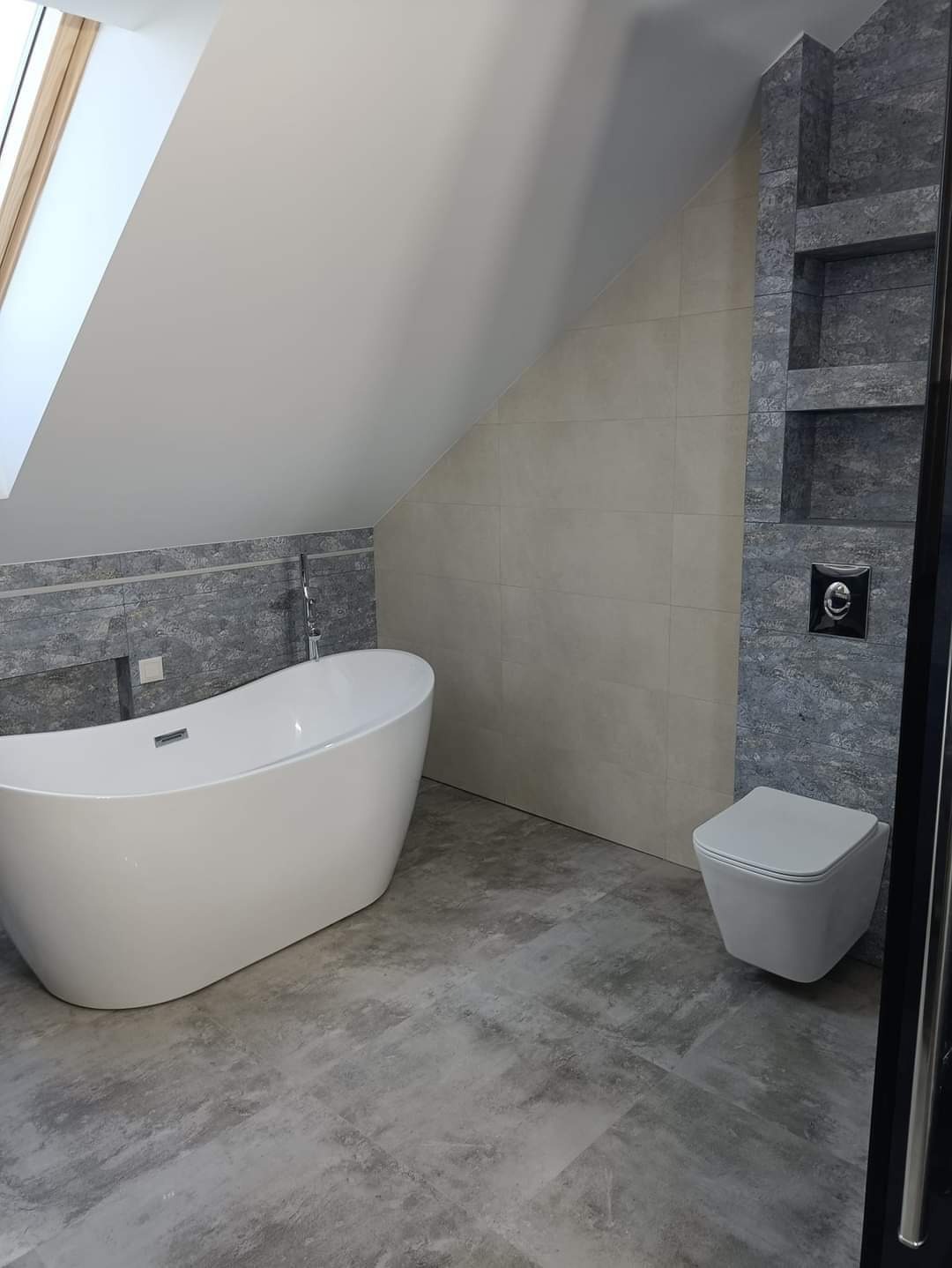 Nowoczesna łazienka na poddaszu z wolnostojącą wanną, podwieszaną toaletą i szarymi płytkami imitującymi beton, oświetlona światłem wpadającym przez okno dachowe.