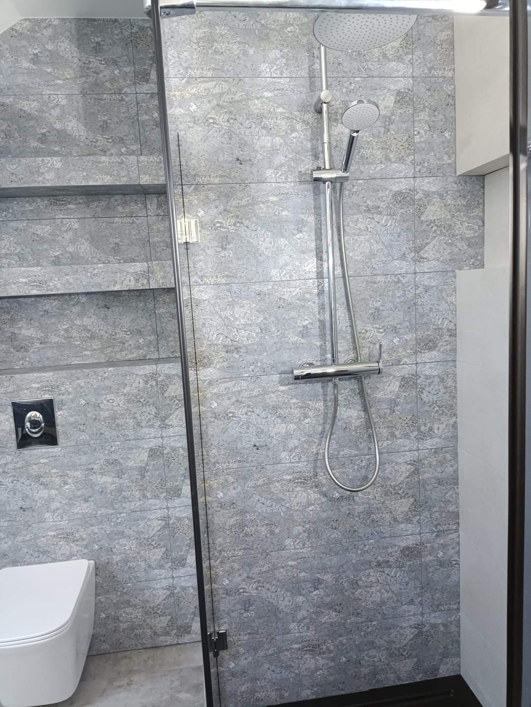 Wykończona łazienka z kabiną prysznicową ze szklanymi drzwiami z czarnymi ramami, deszczownicą i baterią, płyty w odcieniach szarości z wzorem, wisząca toaleta i wnęki półkowe.
