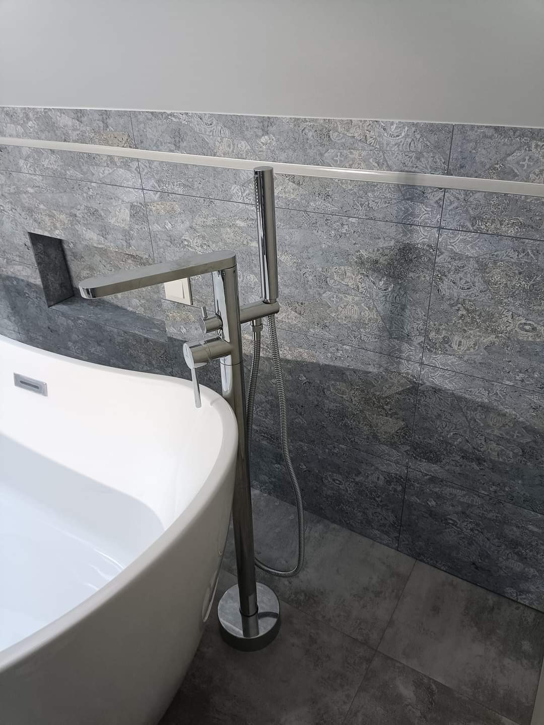 Nowoczesna łazienka z wolnostojącą białą wanną, chromowaną baterią podłogową z prysznicem ręcznym i szarymi płytkami ściennymi z wzorem, uzupełniona listwą dekoracyjną.