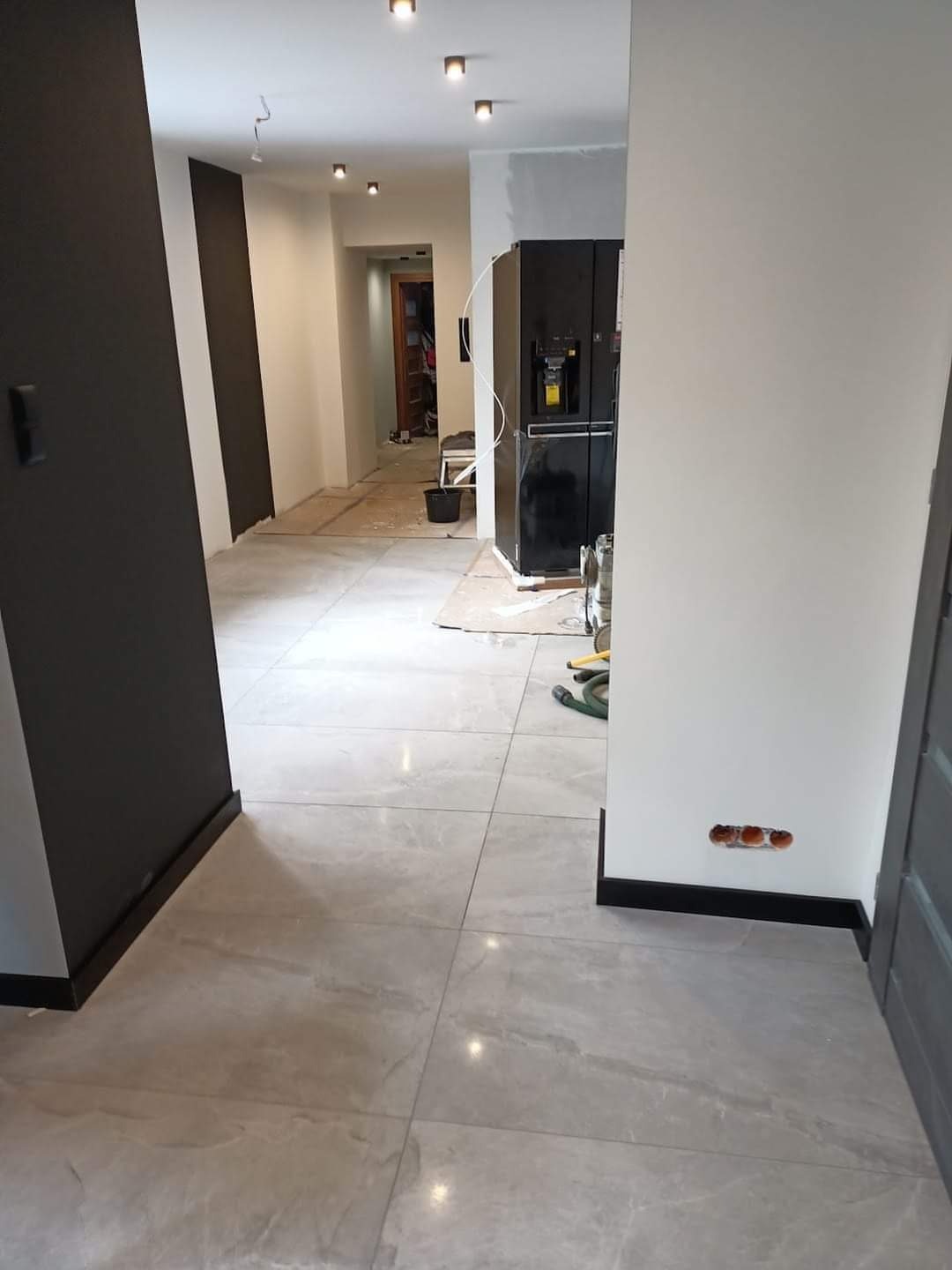 Wnętrze mieszkania w trakcie prac wykończeniowych: szare płytki podłogowe, fragment pomalowanej na szaro ściany, białe ściany, przygotowane puszki elektryczne, w tle sprzęt AGD i otwarte przejście...