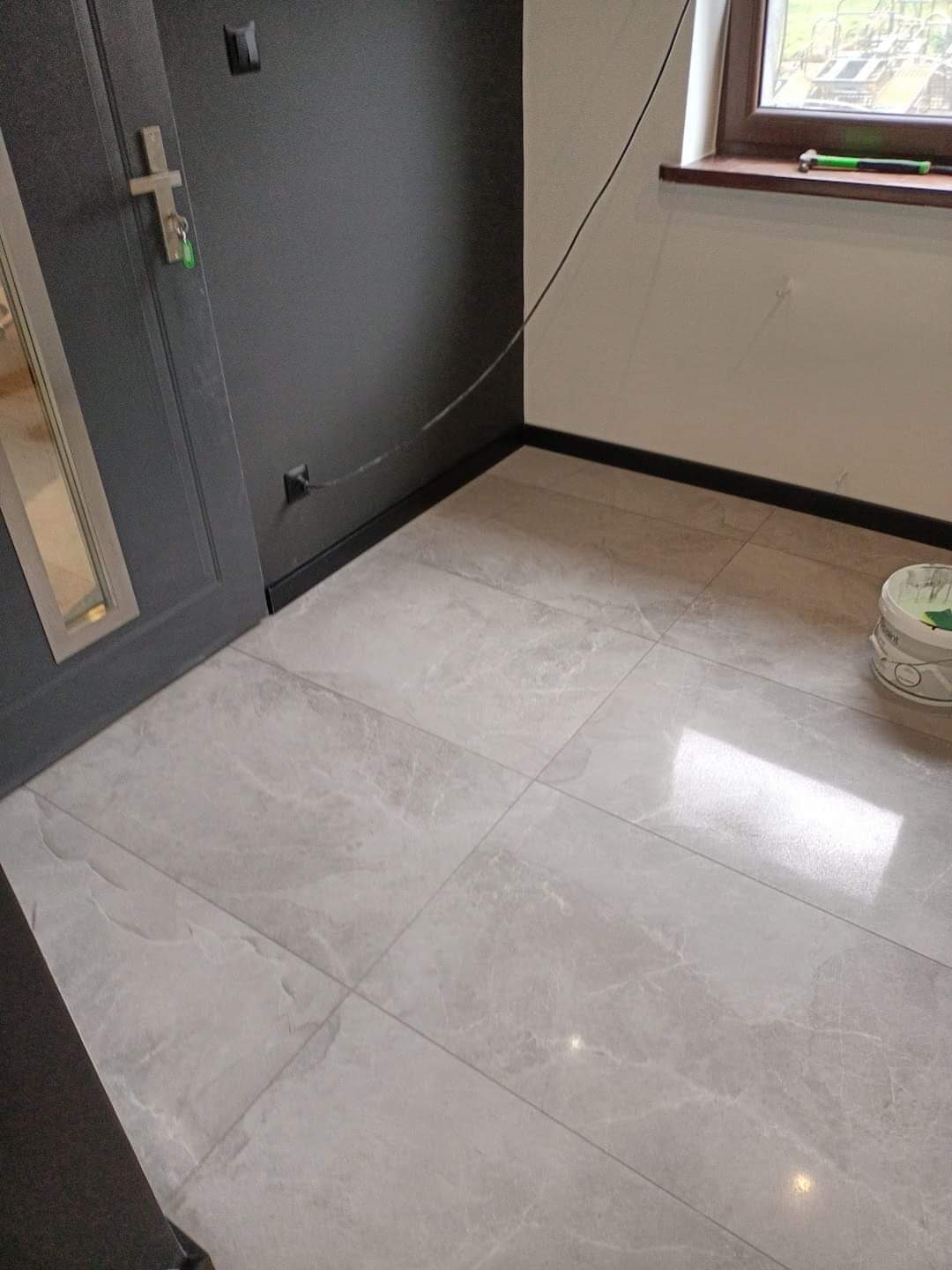 Jasnoszare płytki podłogowe imitujące marmur, ułożone w korytarzu z ciemnymi ścianami i listwami przypodłogowymi, widoczny fragment drzwi i wiadro farby w rogu.