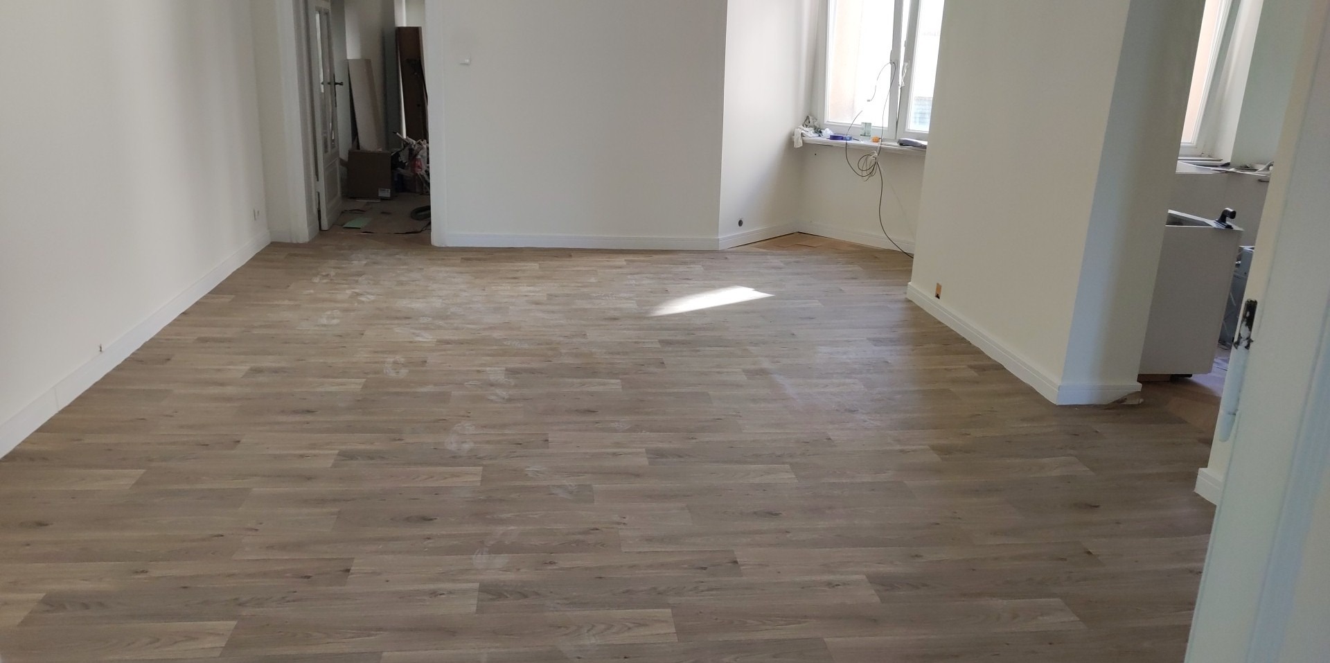 Wykończone, jasne panele podłogowe w nowo pomalowanym pokoju w Szczecinie, widoczne listwy przypodłogowe i fragment okna z parapetem.