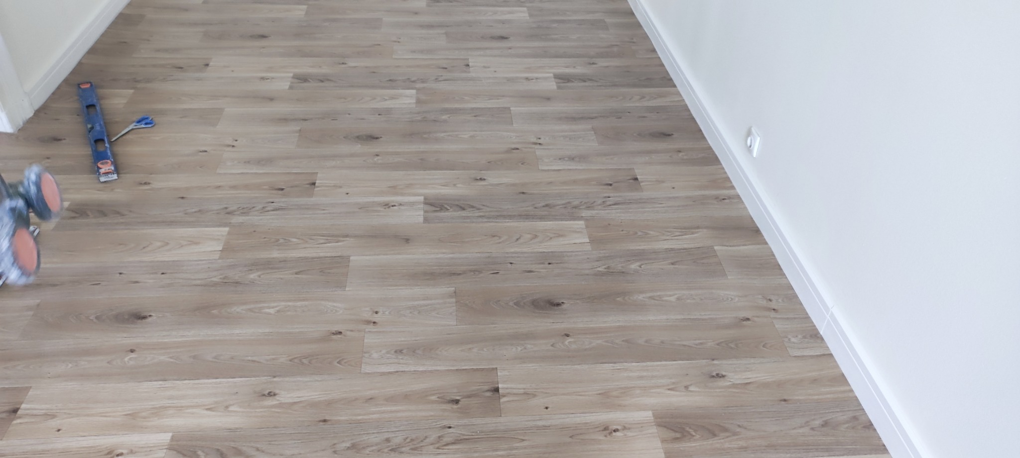 Ułożone panele podłogowe w jasnym odcieniu drewna, widoczna poziomica i nożyczki oraz maszyna do docinania paneli, przy białej ścianie z listwą przypodłogową.