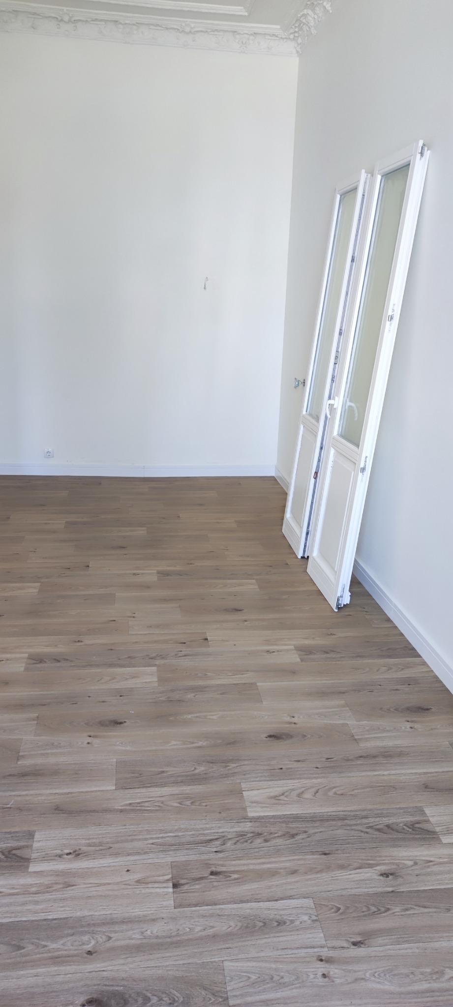 Jasne panele podłogowe z wyraźną strukturą drewna w pustym pomieszczeniu z białymi ścianami i sztukaterią sufitową, oparte o ścianę białe, przeszklone drzwi.