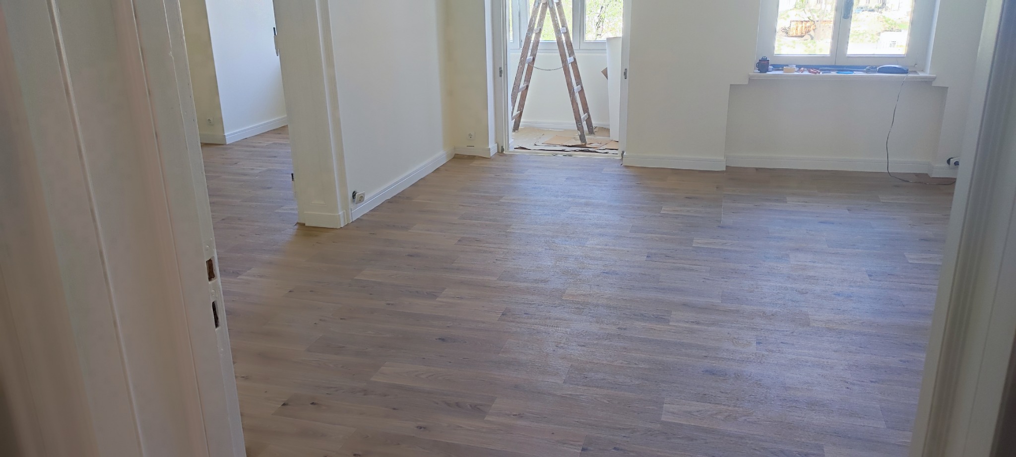 Wykończone wnętrze mieszkania z ułożonymi panelami podłogowymi w odcieniach szarości i beżu, białe ściany i futryny, drabina malarska w tle przy oknie.