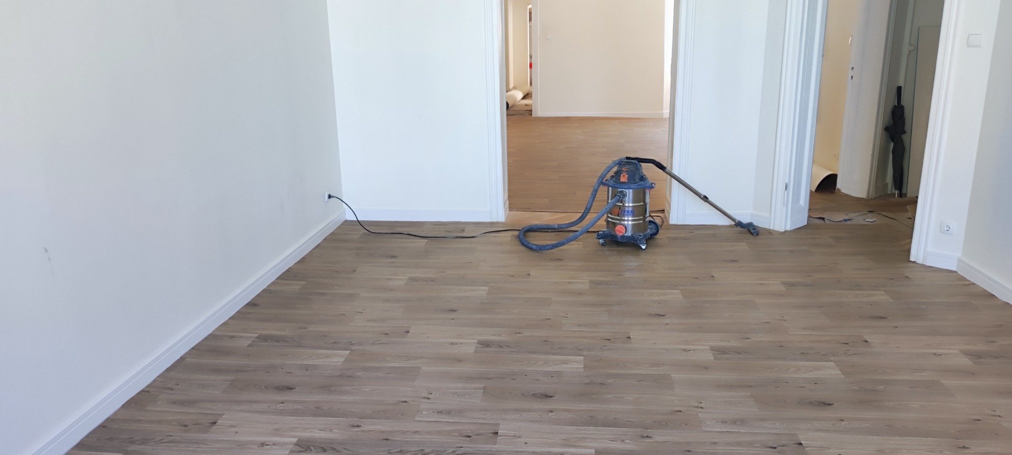 Pokój po ułożeniu paneli podłogowych z widocznym odkurzaczem przemysłowym Dexter i otwartymi drzwiami do kolejnych pomieszczeń z częściowo zabezpieczoną podłogą.