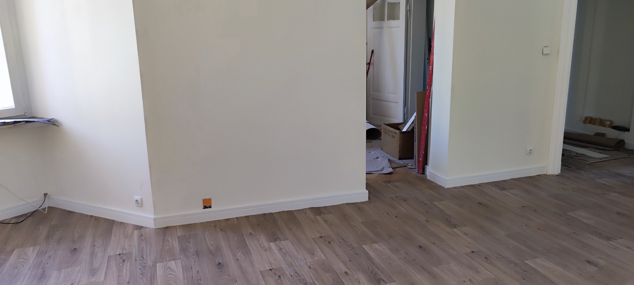 Pokój po remoncie: świeżo ułożone panele podłogowe w odcieniach brązu, białe listwy przypodłogowe, kremowe ściany z widocznym gniazdkiem elektrycznym i otworem po kablach w ścianie, częściowo...