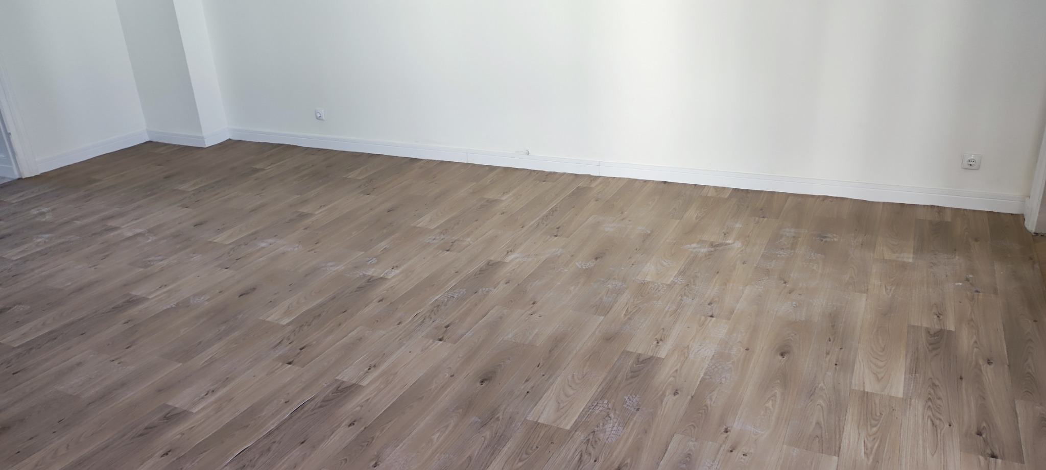 Ułożone panele podłogowe w jasnym odcieniu drewna, widoczne białe listwy przypodłogowe i gniazdko elektryczne na ścianie.