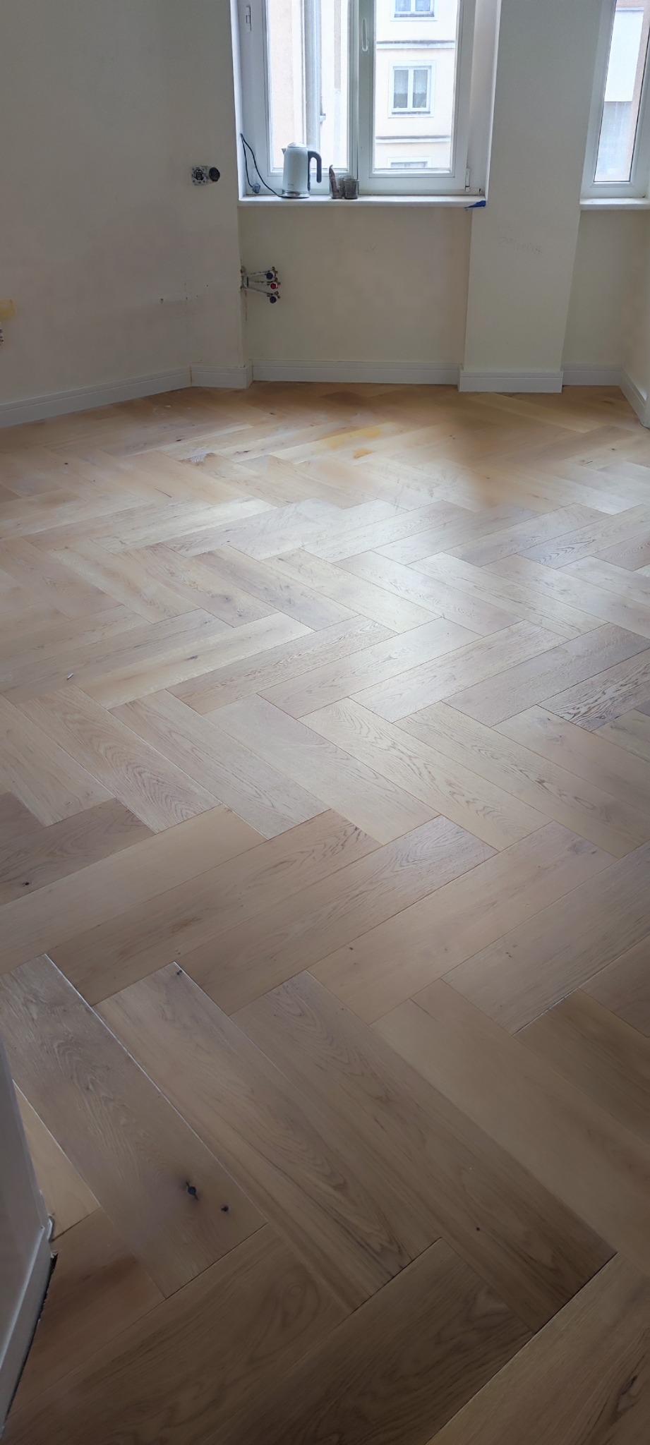 Jasny parkiet ułożony w jodełkę francuską w pustym pokoju z oknem, widoczne ślady po instalacjach na ścianie.