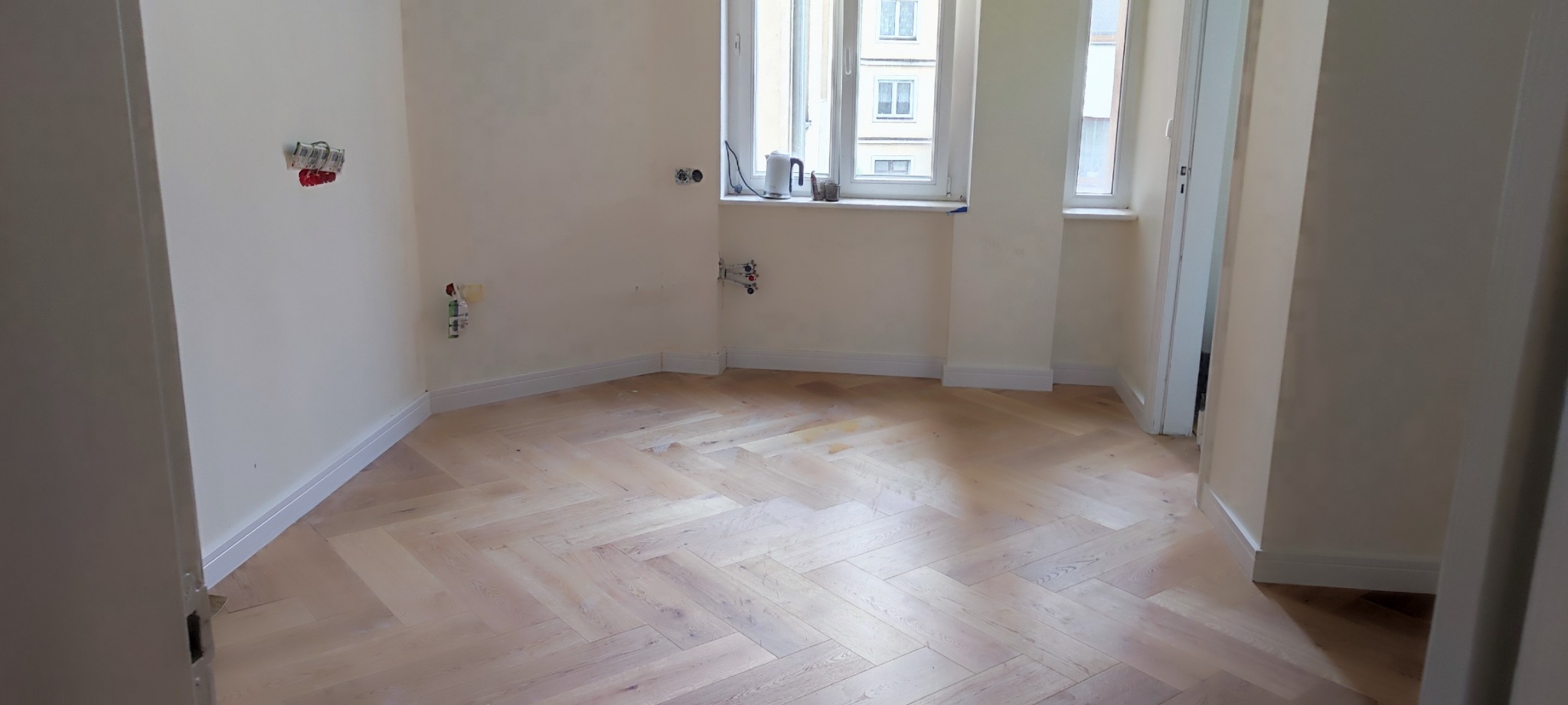 Jasne, świeżo położone panele podłogowe w jodełkę, widoczne przygotowania do instalacji elektrycznej i hydraulicznej na ścianach, surowe wykończenie ścian w pustym pomieszczeniu.