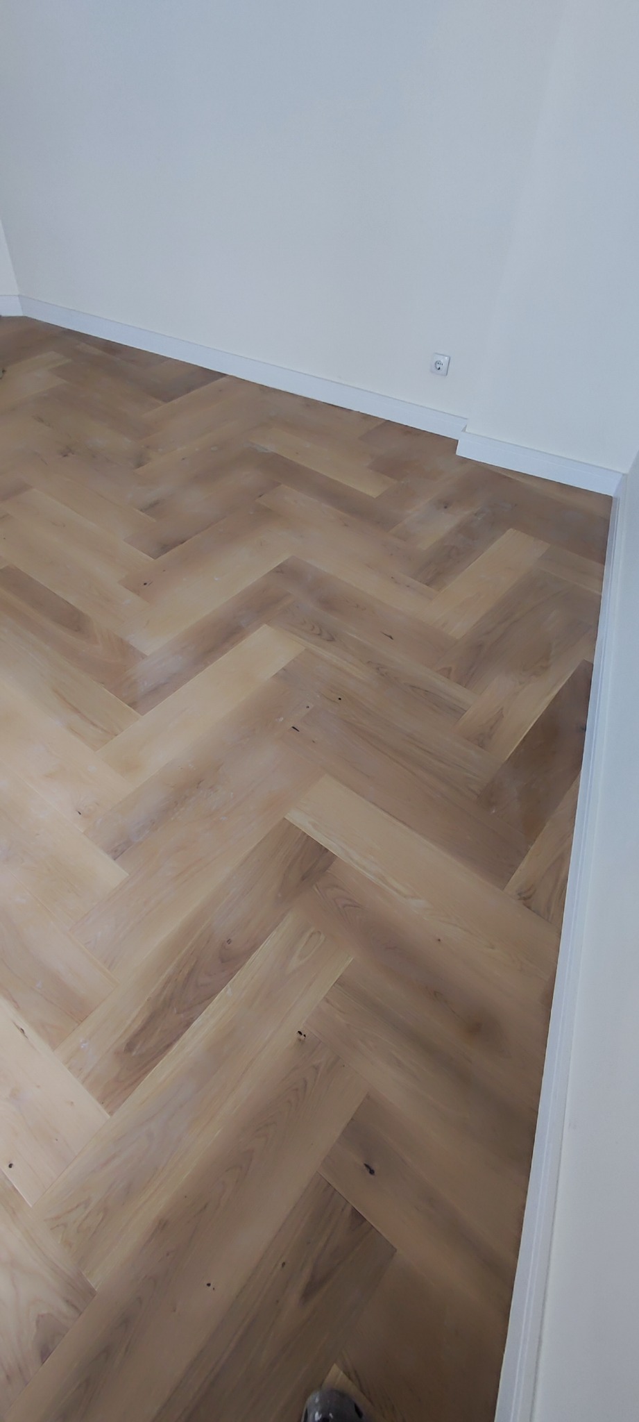 Jasne panele ułożone w jodełkę na podłodze, widoczny fragment białej ściany z listwą przypodłogową i gniazdkiem elektrycznym.