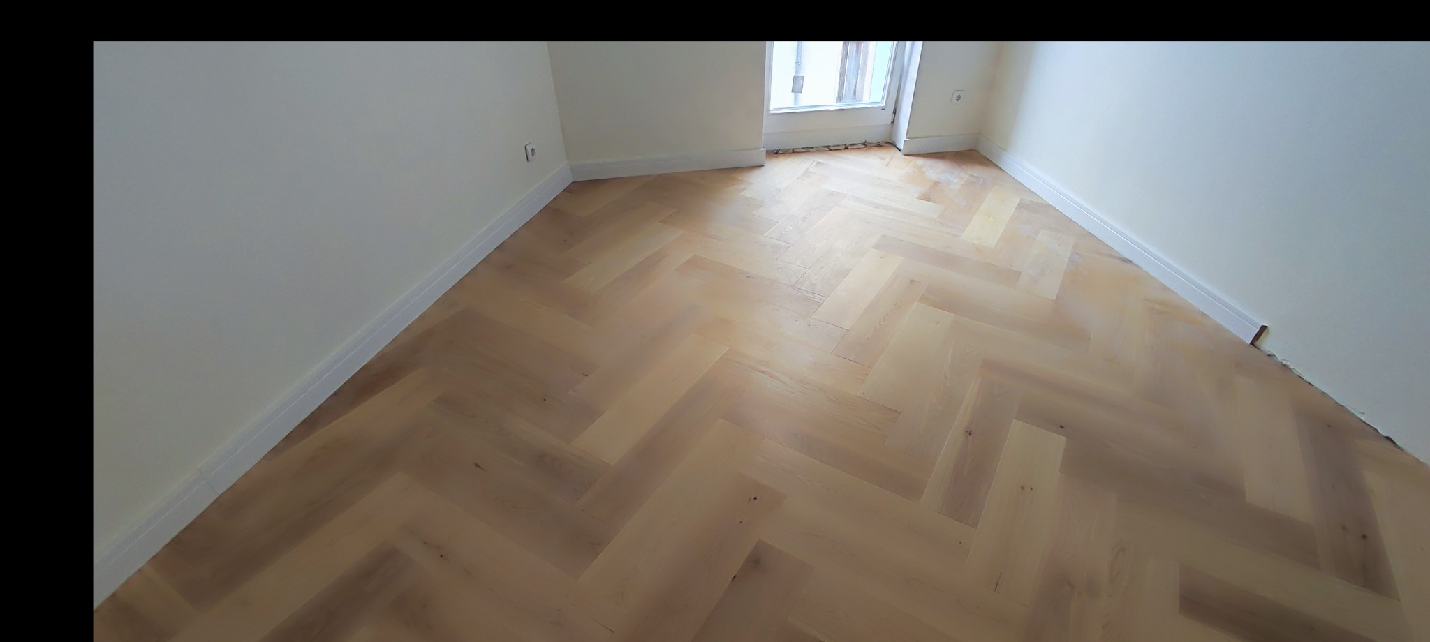 Jasna deska barlinecka ułożona w jodełkę francuską w nowym, jasnym pomieszczeniu z białymi ścianami i listwami przypodłogowymi.