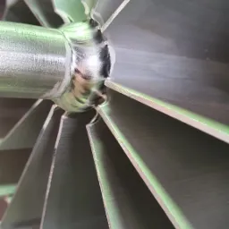 Metalowa turbina z widocznymi spawami łączącymi łopatki z osią, oświetlona z boku, ukazująca detale spoin.