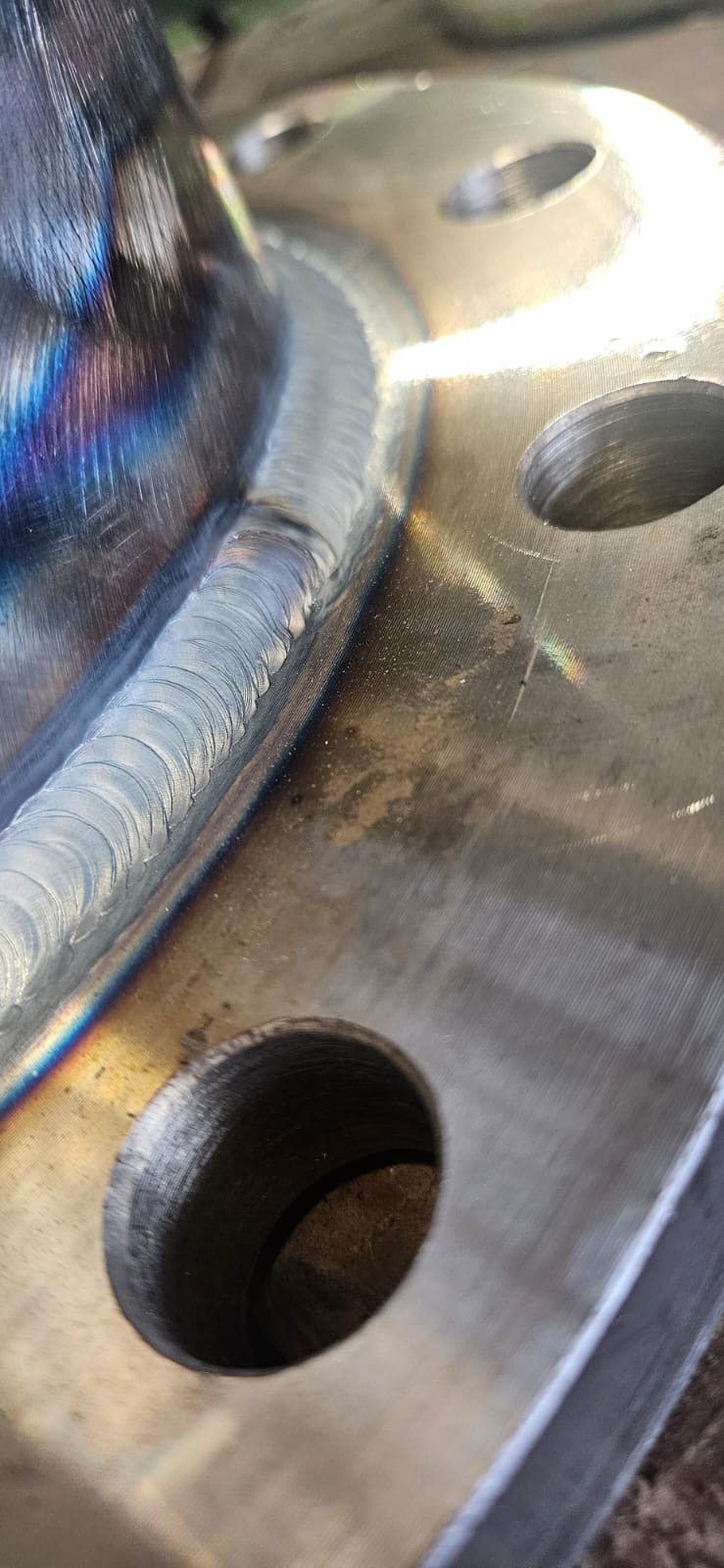 Szczegółowe zbliżenie na spawany element metalowy z widocznymi tęczowymi przebarwieniami po spawaniu i otworami. Precyzyjne spawy, gładka powierzchnia metalu.