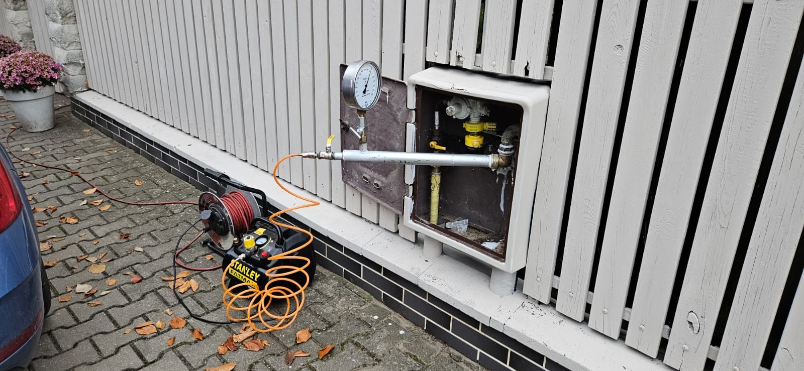 Próba szczelności instalacji gazowej: manometr podłączony do skrzynki gazowej na elewacji budynku, widoczny kompresor Stanley Fatmax i zwinięty przewód.