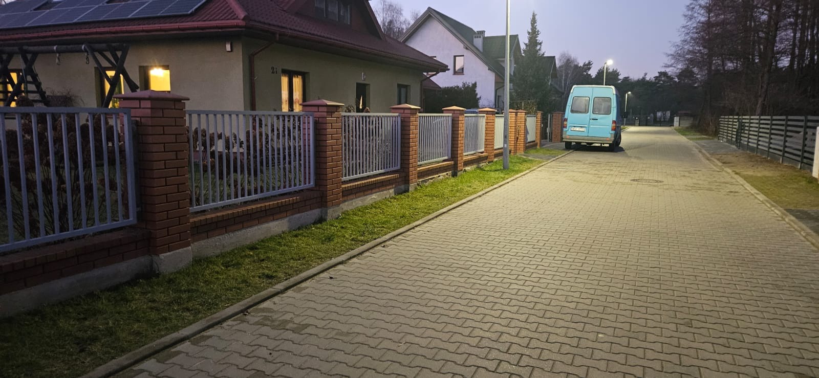 Nowoczesne ogrodzenie metalowe z ceglanymi słupkami, widok z ulicy w szarym świetle. Błękitny bus zaparkowany w tle, podkreślający realizację.