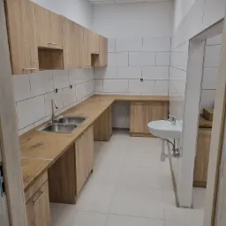 Jasna kuchnia z drewnianymi szafkami i białymi płytkami, widoczny zlew dwukomorowy i umywalka ceramiczna, minimalizm.