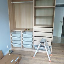 Piotr Bogdański - Montaż jasnej, drewnianej szafy z szufladami i koszami; widoczna drabina, wkrętarka i kartony z elementami na podłodze.