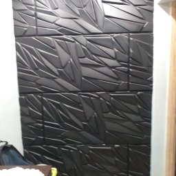 Piotr Bogdański - Czarny panel ścienny 3D z geometrycznym wzorem, ułożony z kilku kwadratowych płyt, widoczny fragment białej ściany i futryny drzwi.