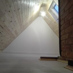 Piotr Bogdański - Wykończone poddasze z drewnianą boazerią na skosach, białą ścianą szczytową, oknem dachowym i fragmentem ceglanego komina.
