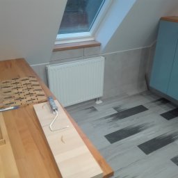 Piotr Bogdański - Przygotowania do malowania w pokoju na poddaszu: wałek malarski z niebieską rączką leży na drewnianym blacie obok pojemnika na farbę, widoczny grzejnik pod oknem dachowym i szafka w odcieniach...