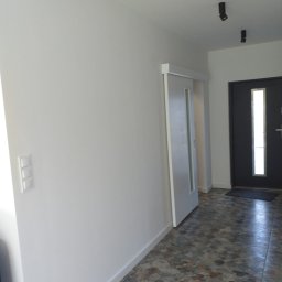 Piotr Bogdański - Wykończony korytarz w Szczecinie z przesuwnymi drzwiami, białymi ścianami z widocznymi włącznikami, ciemnymi punktowymi lampami sufitowymi i podłogą w patchworkowym stylu.