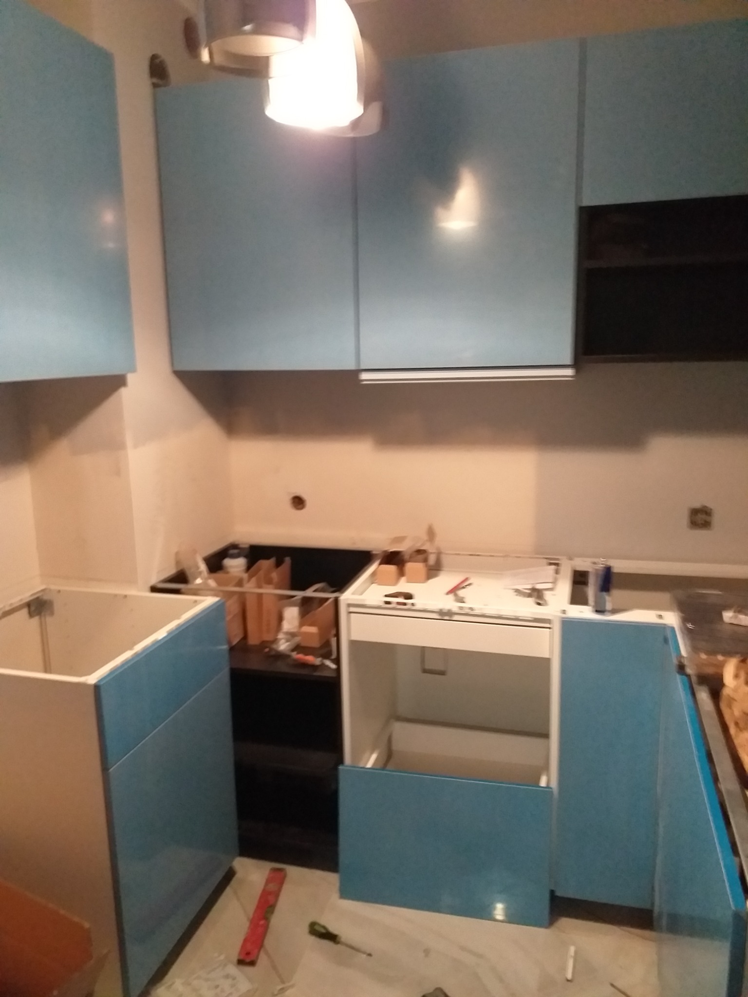 Niedokończony montaż kuchni: błękitne szafki wiszące i stojące, widoczne narzędzia, poziomica i śrubokręt na podłodze, brakujące elementy blatu i wykończenia.