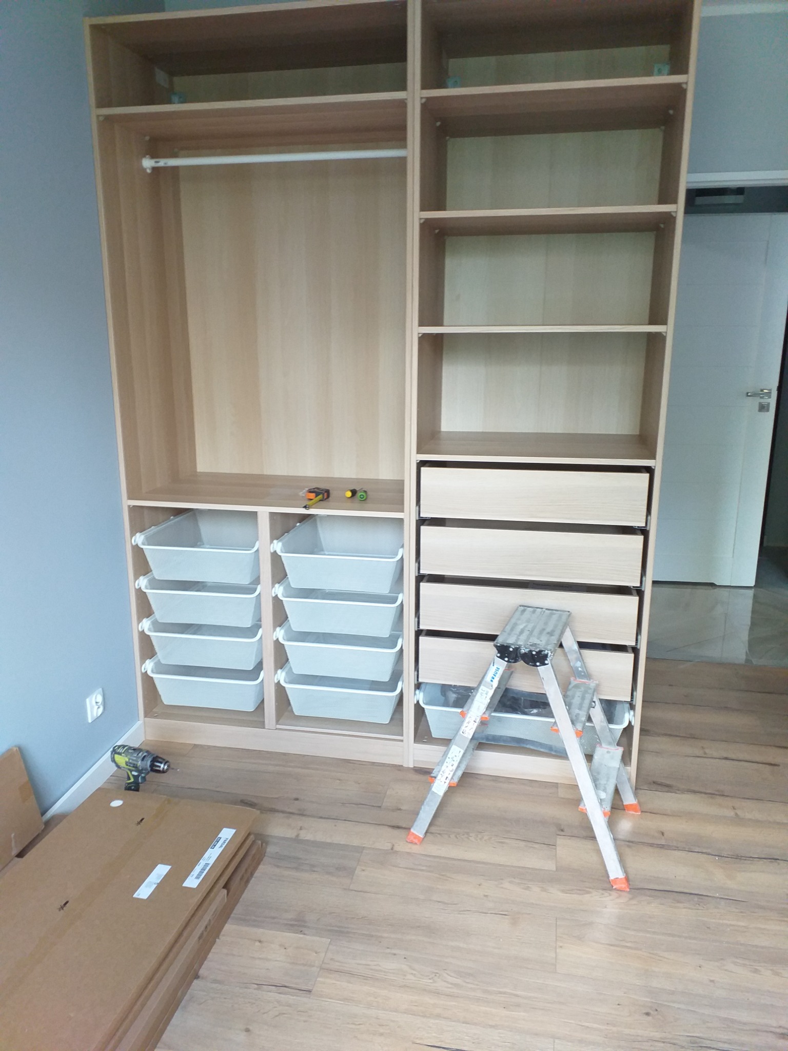 Montaż jasnej, drewnianej szafy z szufladami i koszami; widoczna drabina, wkrętarka i kartony z elementami na podłodze.