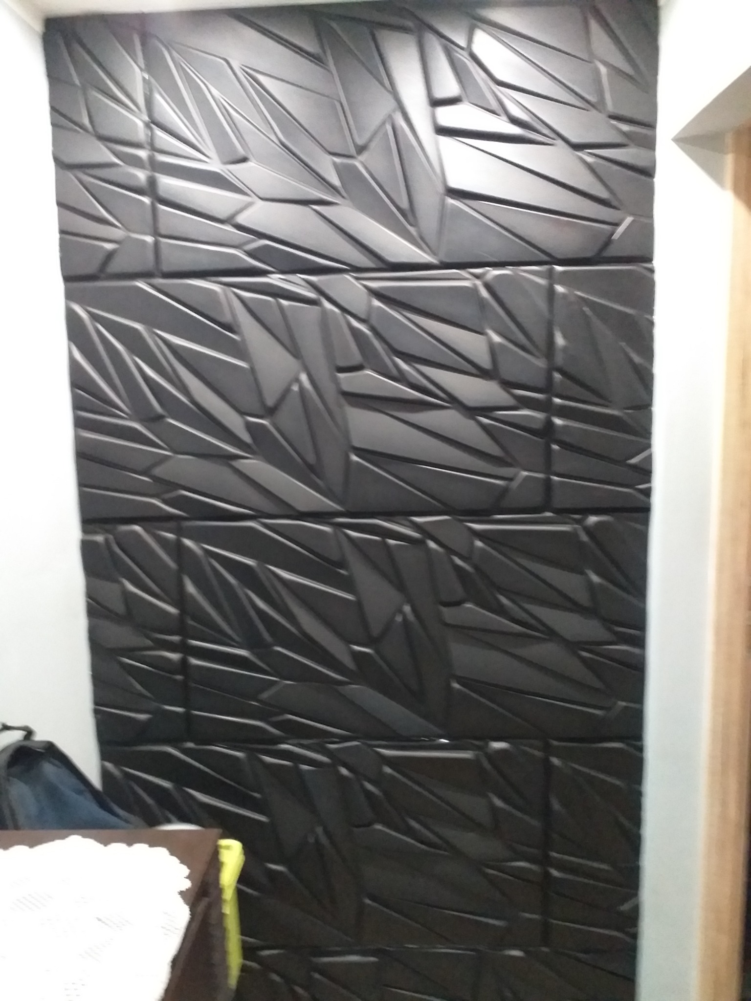 Czarny panel ścienny 3D z geometrycznym wzorem, ułożony z kilku kwadratowych płyt, widoczny fragment białej ściany i futryny drzwi.