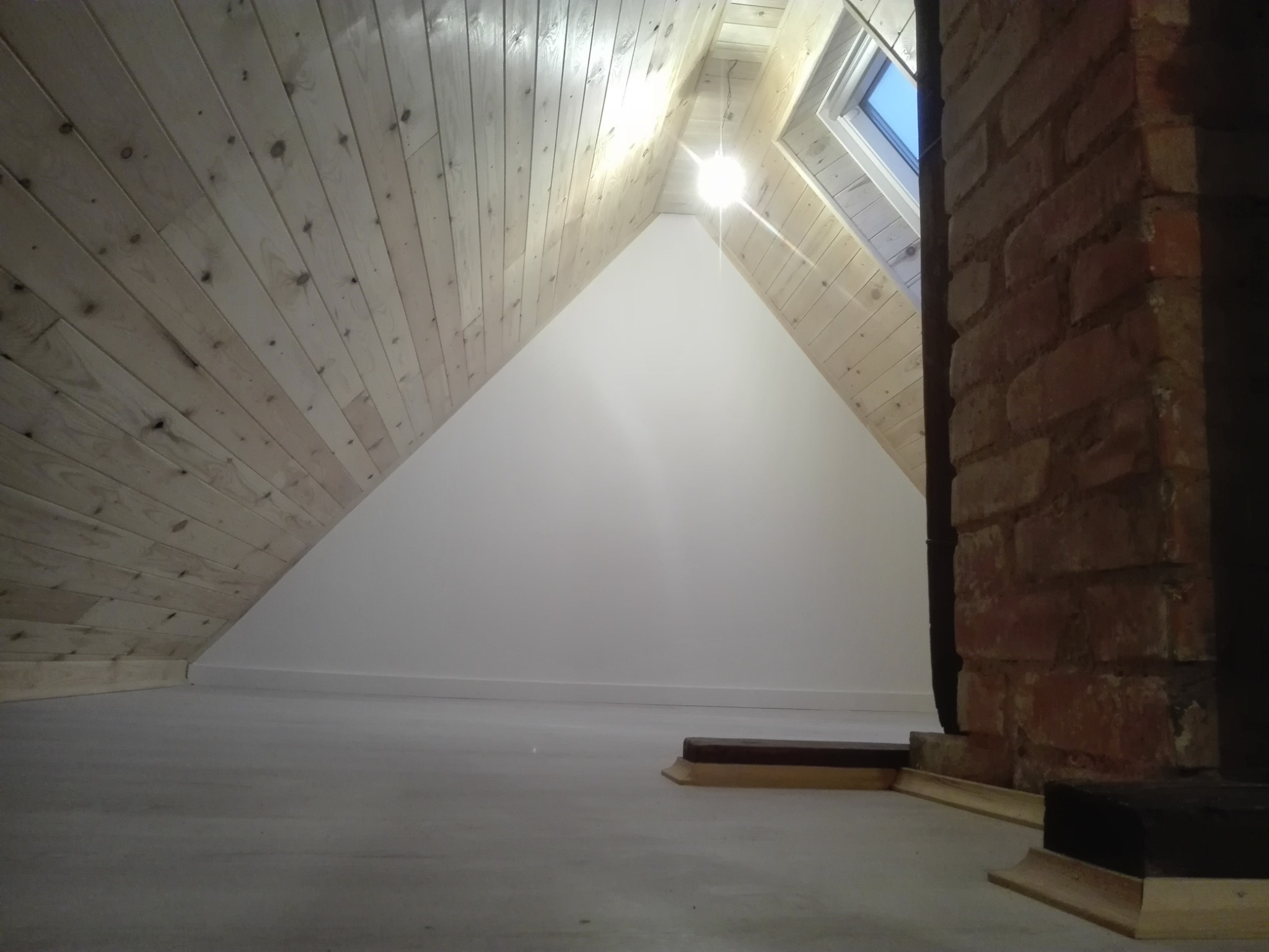 Wykończone poddasze z drewnianą boazerią na skosach, białą ścianą szczytową, oknem dachowym i fragmentem ceglanego komina.