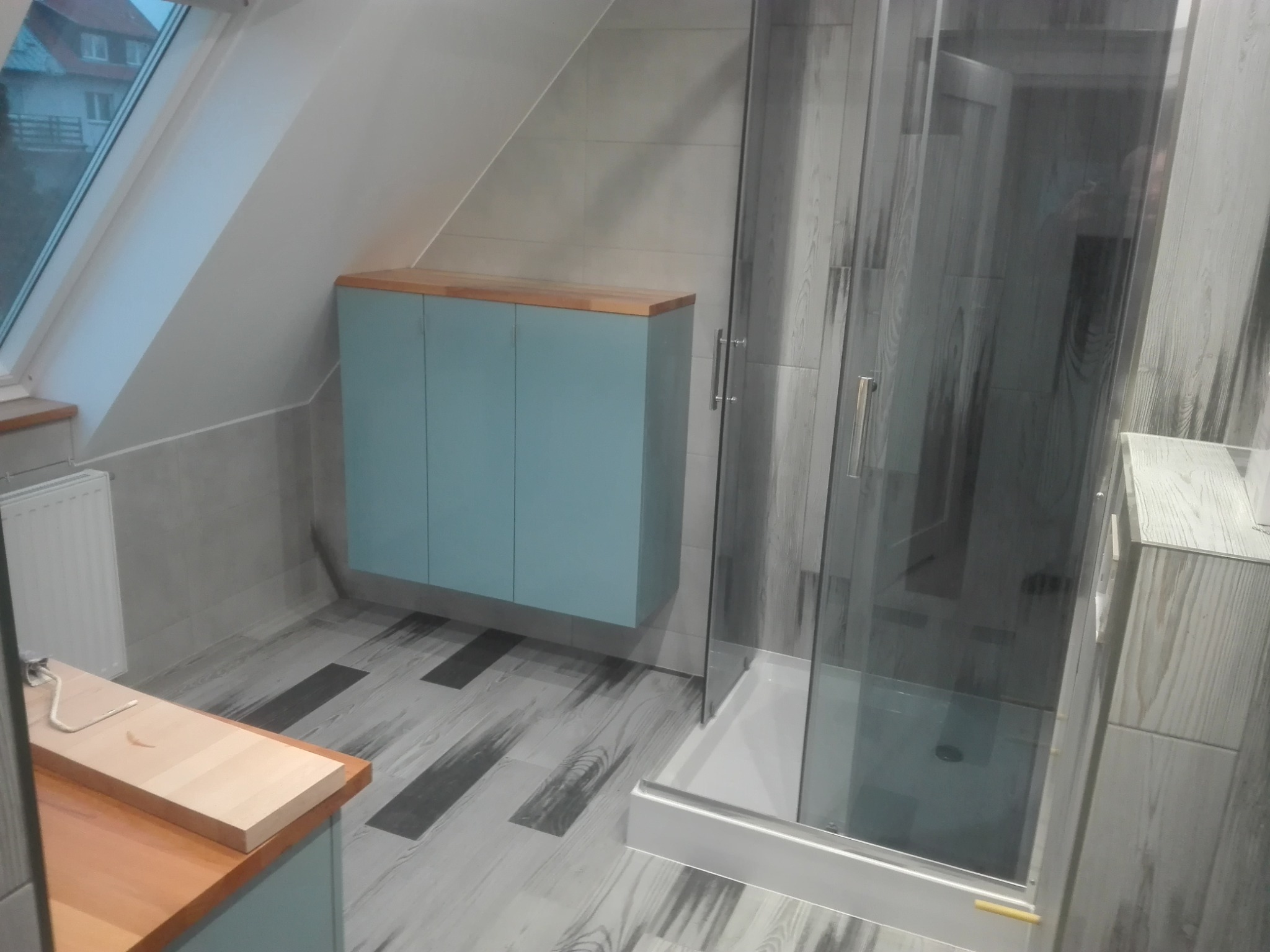 Wykończona łazienka na poddaszu ze skośnym sufitem: kabina prysznicowa z przezroczystego szkła, szafka wisząca w kolorze błękitnym z drewnianym blatem, podłoga z płytek imitujących drewno...