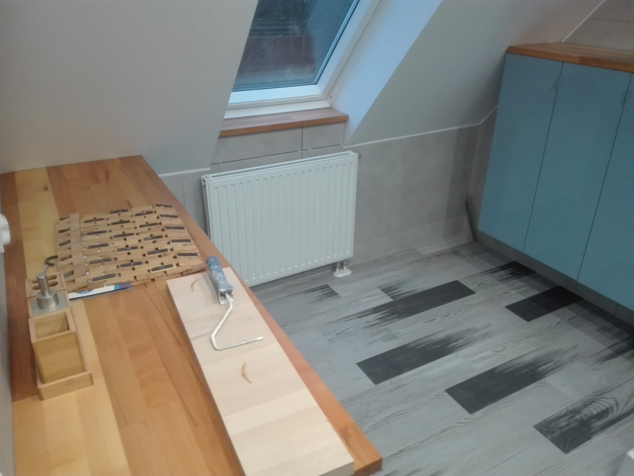 Przygotowania do malowania w pokoju na poddaszu: wałek malarski z niebieską rączką leży na drewnianym blacie obok pojemnika na farbę, widoczny grzejnik pod oknem dachowym i szafka w odcieniach...