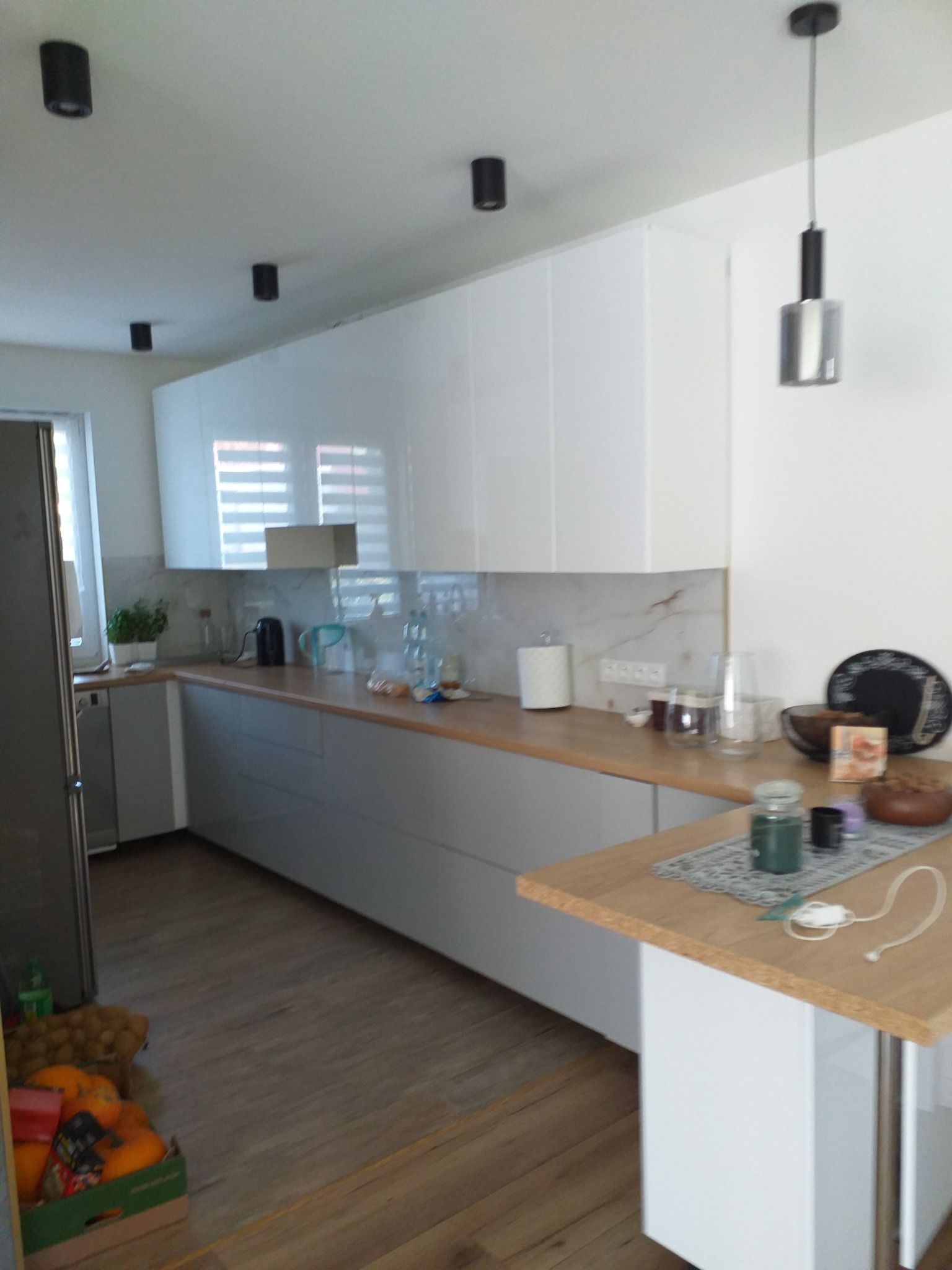 Nowoczesna kuchnia z białymi szafkami, drewnianym blatem i wyspą, oświetlona punktowymi lampami sufitowymi i wiszącą lampą, z widocznymi akcesoriami kuchennymi i dekoracjami.