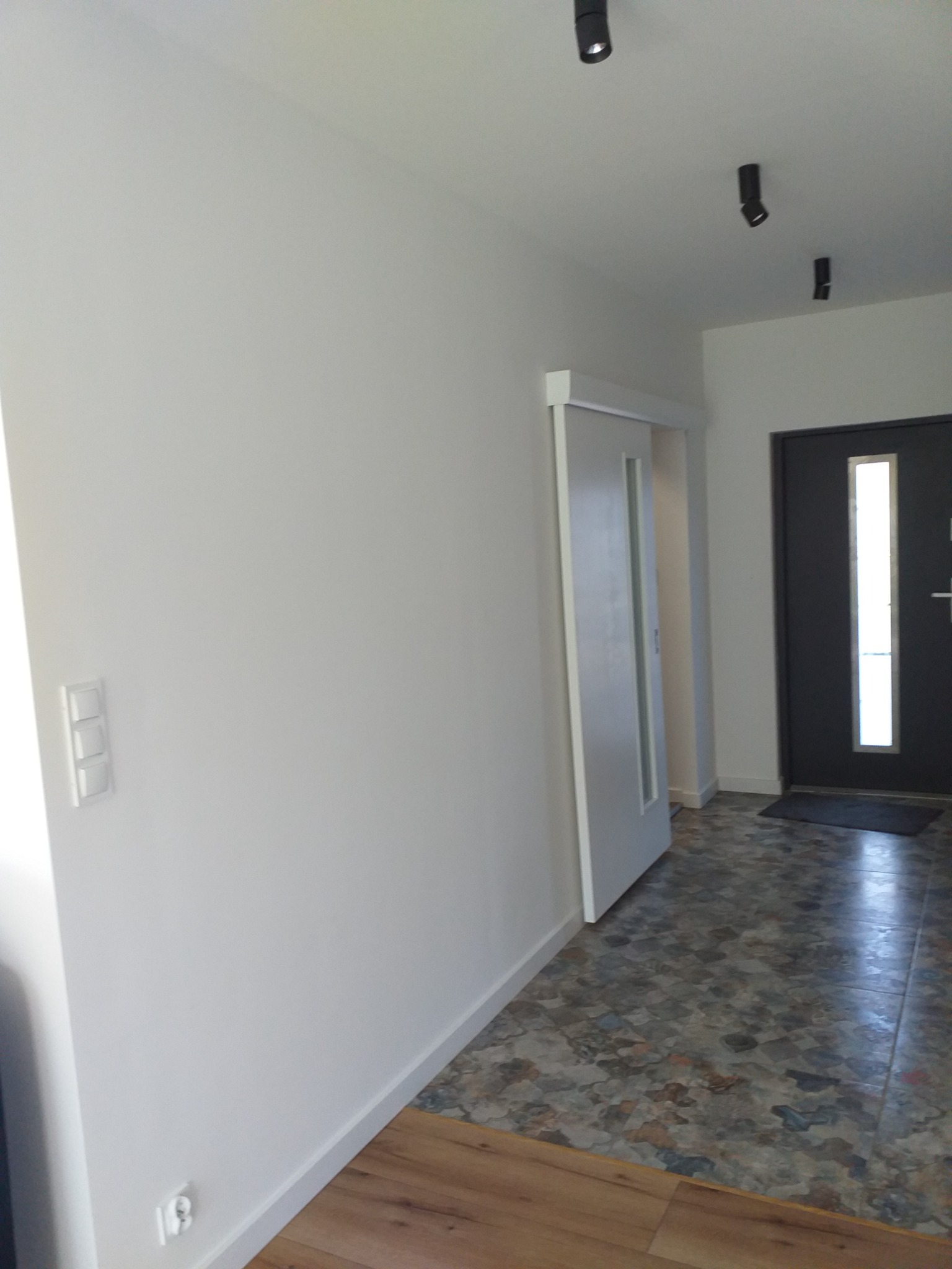 Wykończony korytarz w Szczecinie z przesuwnymi drzwiami, białymi ścianami z widocznymi włącznikami, ciemnymi punktowymi lampami sufitowymi i podłogą w patchworkowym stylu.