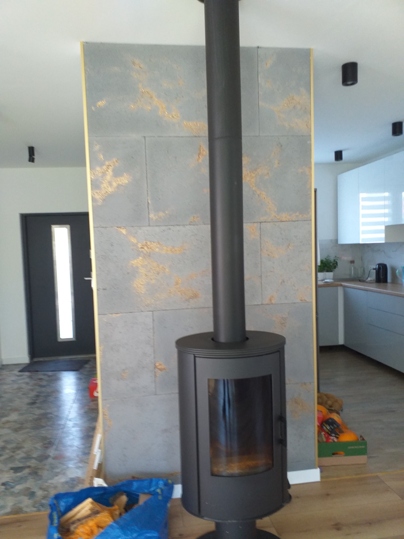 Nowoczesny piec wolnostojący z długą rurą kominową ustawiony na tle ściany z szarej płyty imitującej kamień z złotymi akcentami, widoczne elementy kuchni i drzwi wejściowe.