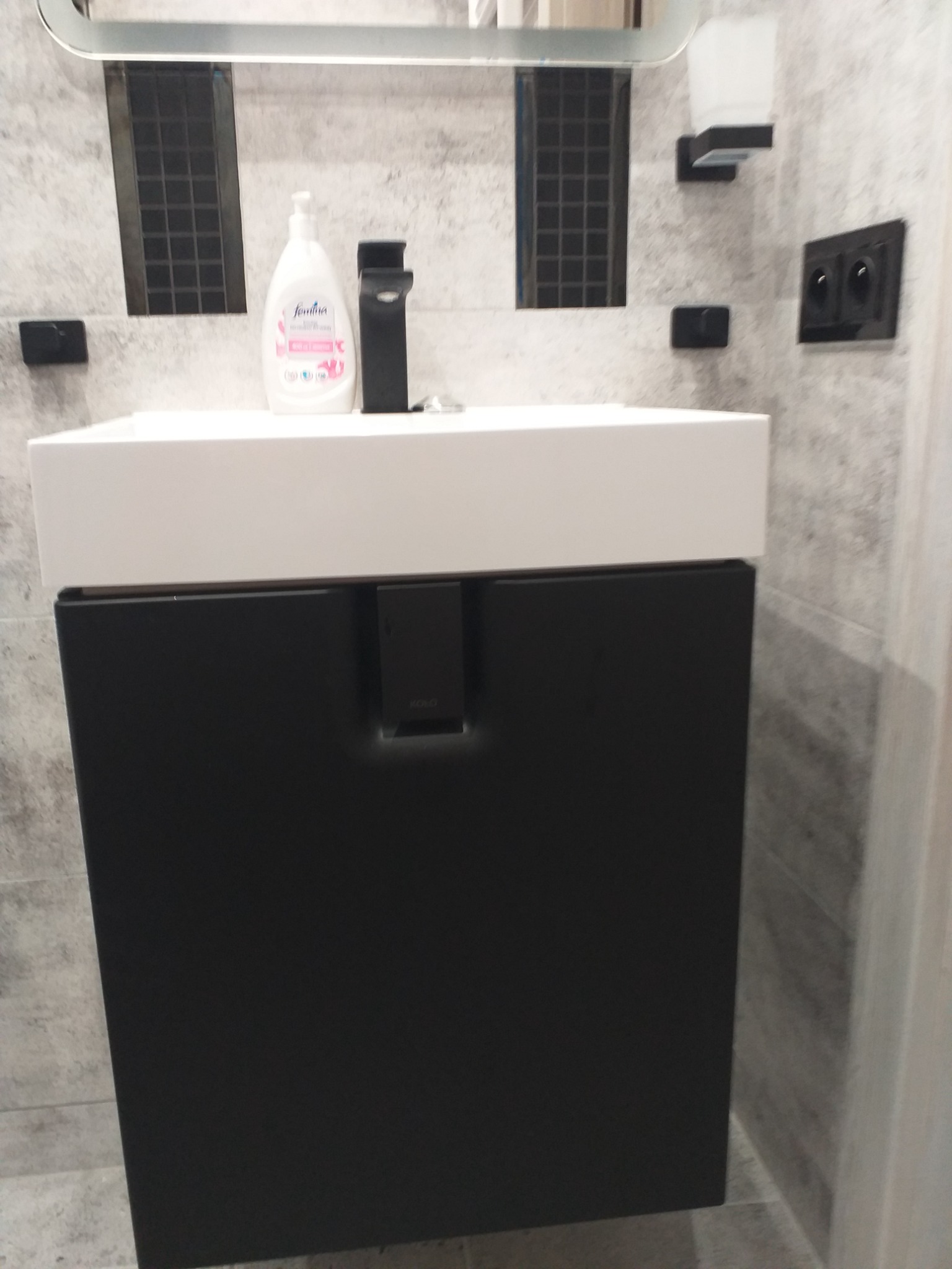 Nowoczesna łazienka z umywalką nablatową na czarnej szafce, czarna bateria umywalkowa, szara płytka imitująca beton, czarne akcesoria łazienkowe i dozownik mydła.