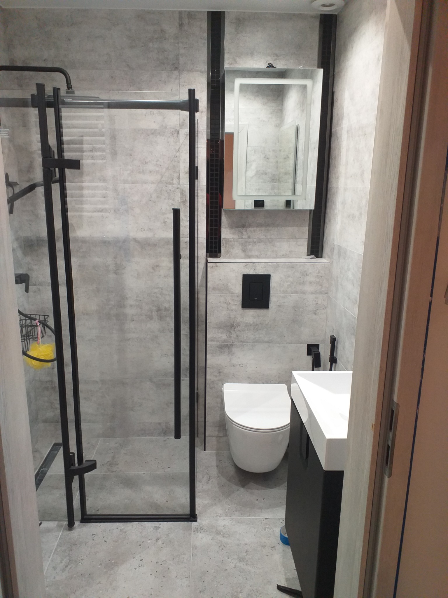 Nowoczesna łazienka w szarościach z kabiną prysznicową z czarnymi ramami, podwieszaną toaletą i umywalką na szafce, oświetlone lustro.