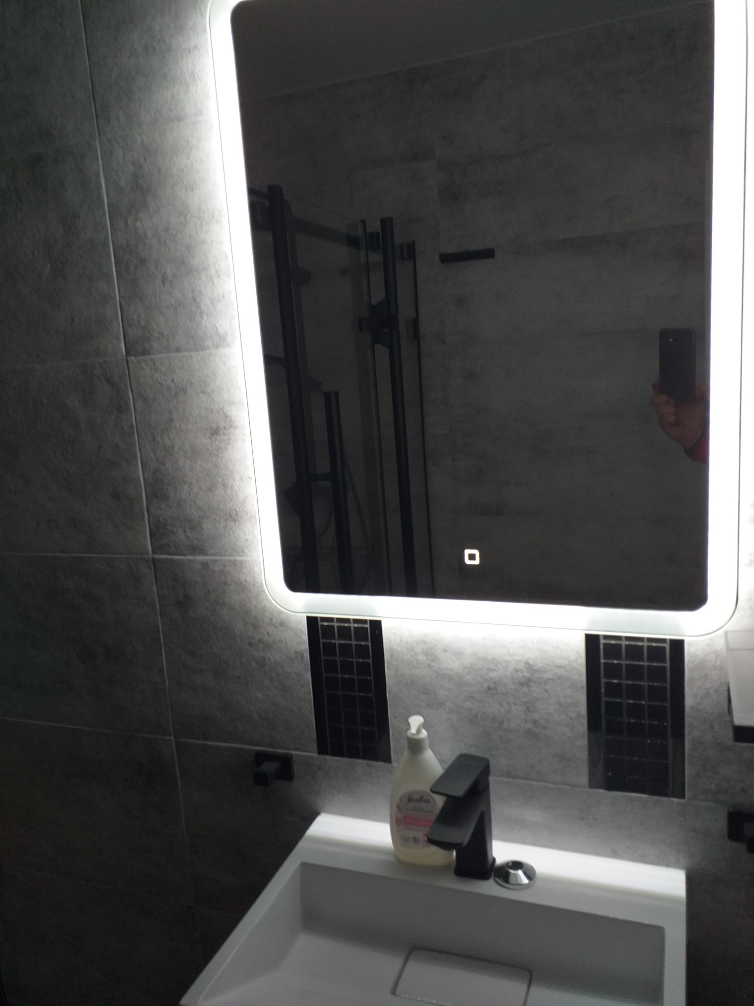 Nowoczesna łazienka z szarymi płytkami, podświetlanym lustrem, czarną armaturą i dozownikiem mydła na białej umywalce; widoczne odbicie osoby robiącej zdjęcie.