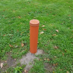 Pomarańczowa rura studni wystająca z trawnika, widoczne liście na trawie i ziemia wokół rury.