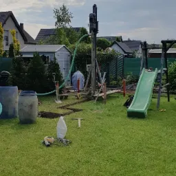 Wiercenie studni głębinowej w ogrodzie, widoczna wiertnica, węże, narzędzia, wykopana bruzda w trawie, w tle dom i plac zabaw z zieloną zjeżdżalnią.