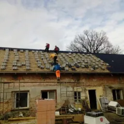 Ekipa dekarzy na dachu starego, ceglanego budynku, częściowo pokrytym nową, czarną dachówką, z widocznymi rusztowaniami i stertą cegieł na ziemi.