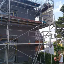 Dom w trakcie ocieplania: czarne płyty izolacyjne przymocowane do ściany, rusztowanie z siatką ochronną, pracownik przygotowuje przewód elektryczny.