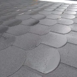 Szczegółowe ujęcie nowo położonego, grafitowego gontu bitumicznego na dachu, z widoczną fakturą i częściowym połyskiem, oraz fragmentem pracownika w tle.