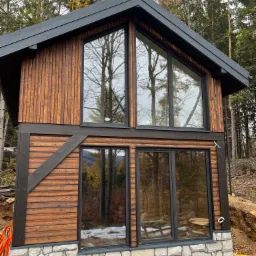 Dwukondygnacyjny budynek z drewnianą elewacją i dużymi oknami, posadowiony na fundamencie z kamiennej okładziny, otoczony drzewami.