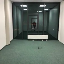 Puste biuro z zieloną wykładziną, grzejnikiem pod oknem i widokiem na korytarz przez szklaną ścianę. W odbiciu okna widoczna sylwetka osoby.