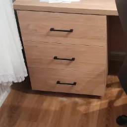 Biurko z trzema szufladami o drewnianym wykończeniu i czarnymi uchwytami, ustawione na panelach podłogowych, obok grzejnika z firanką i czarnym krzesłem biurowym.