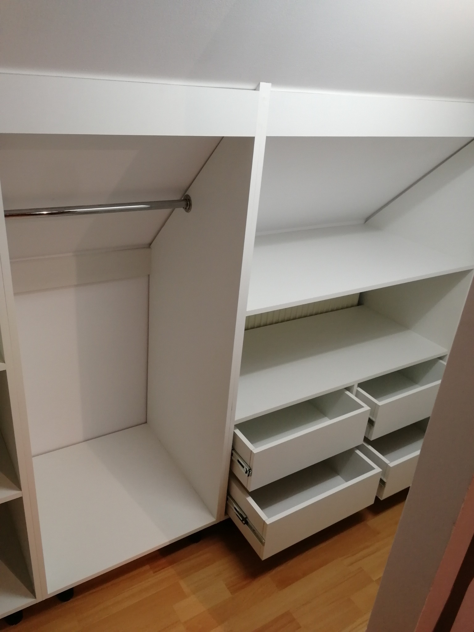 Wnętrze białej szafy na wymiar z drążkiem na ubrania i wysuwanymi szufladami, zamontowanej na poddaszu ze skośnym sufitem.
