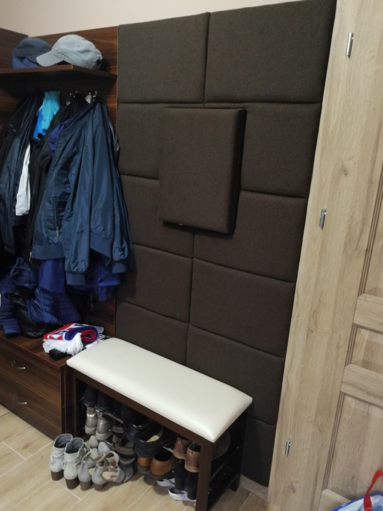 Panel ścienny z brązowych kwadratów, po lewej szafka z ubraniami i butami, po prawej drzwi z jasnego drewna, całość w przedpokoju.